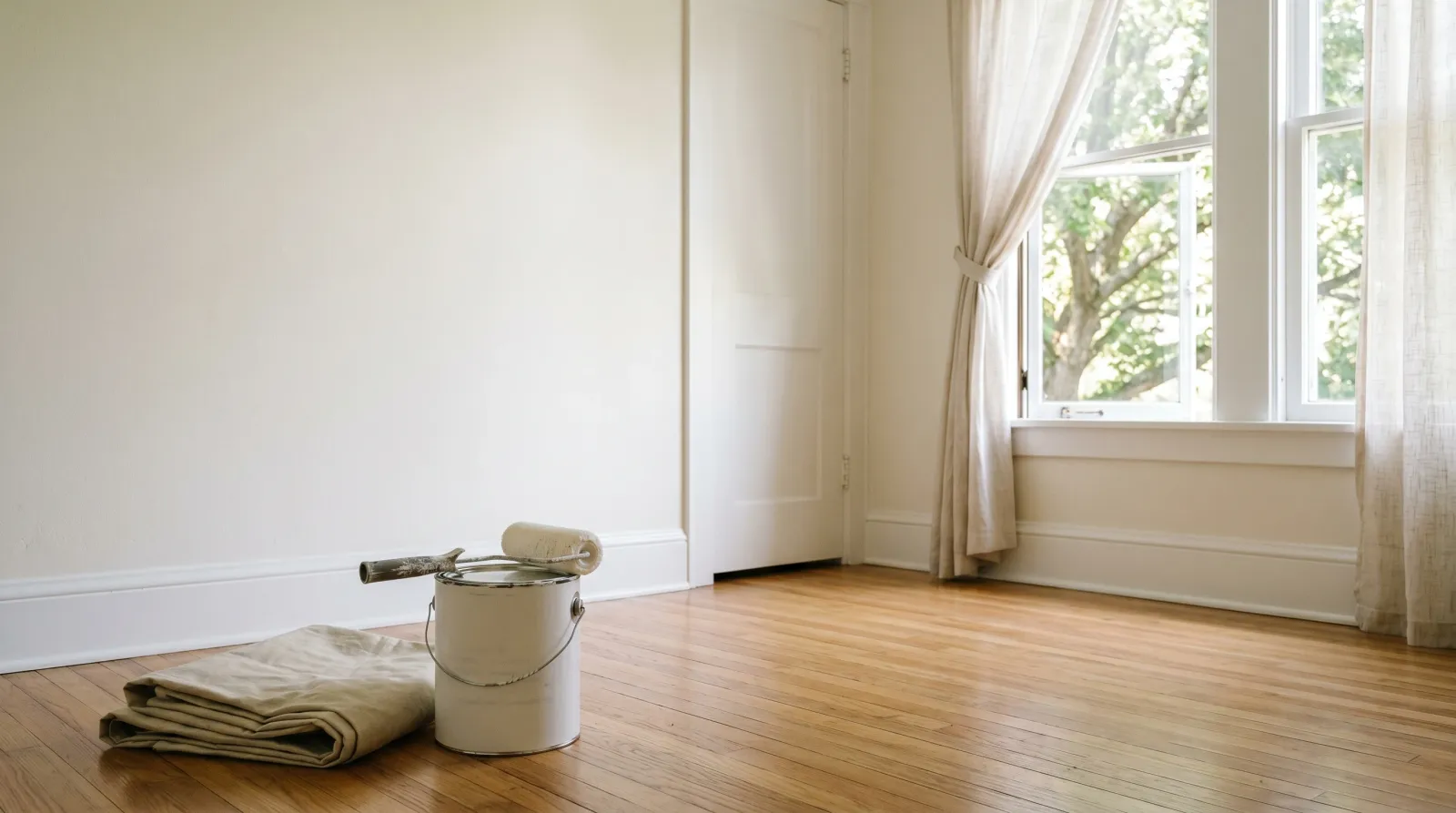 Freshly repainted main-floor bedroom with open windows, a sealed paint can on a drop cloth, late-morning daylight raking across an eggshell wall