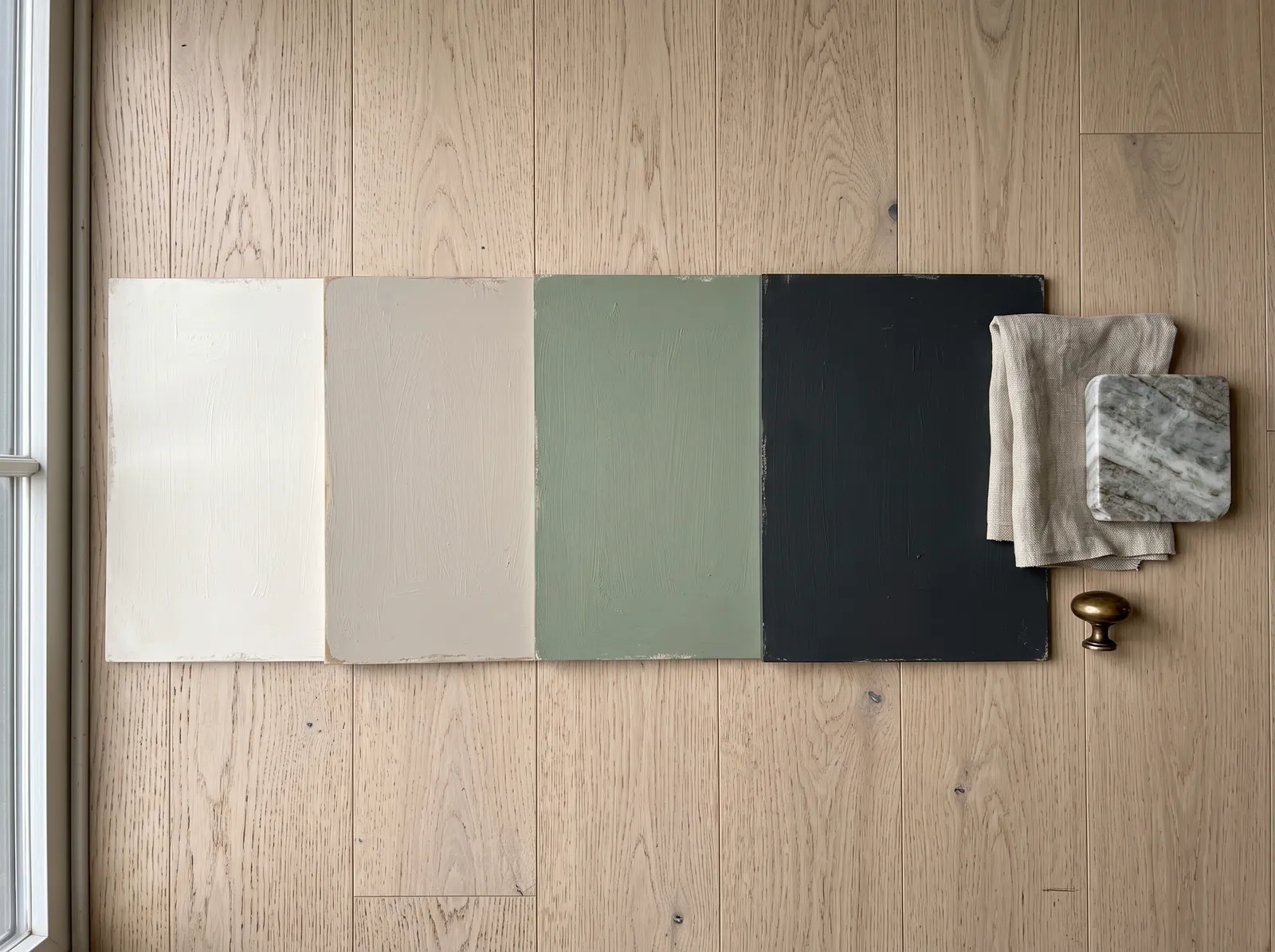 Four painted sample boards laid on an oak floor beside a linen swatch, a brass knob, and a stone countertop offcut