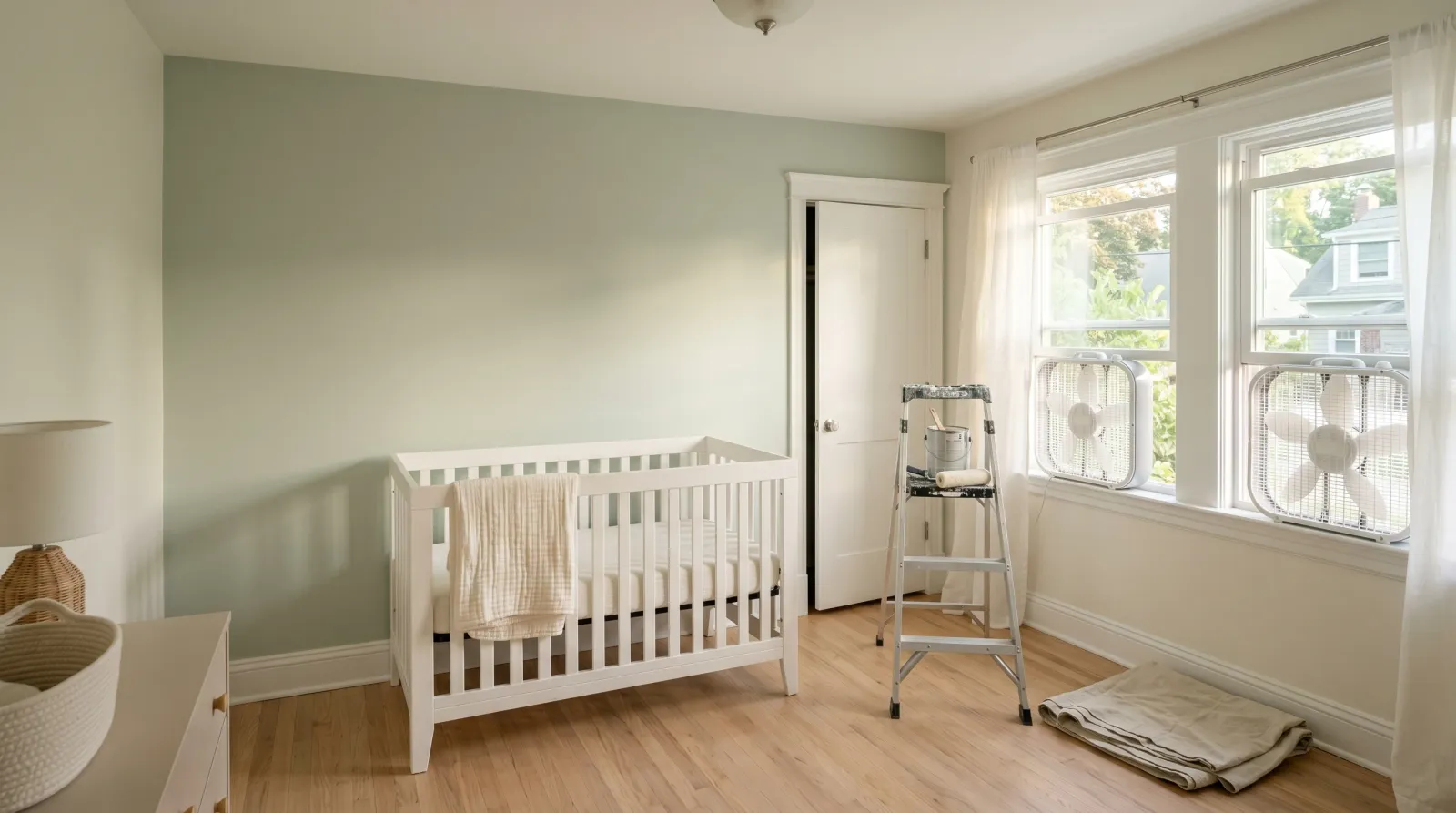 Freshly painted nursery with soft white walls, a pale sage accent wall, white crib, and box fans in the windows